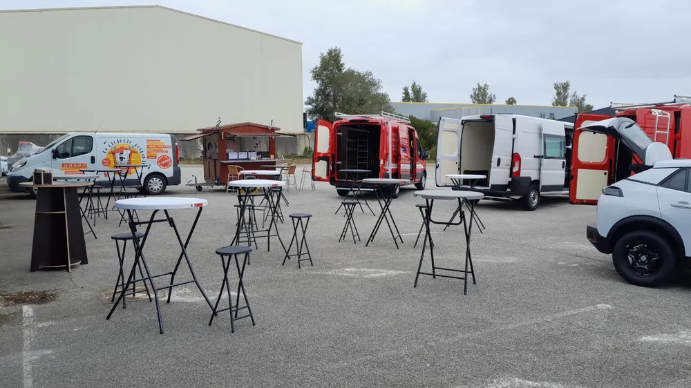 Mange-debout et tabourets installés devant un bâtiment d'entreprise, avec la roulotte en bois et le camion de La Crêperie Roulante