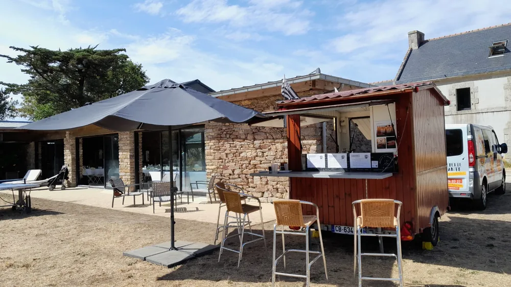 Roulotte à crêpes, parasol et tabourets hauts devant une maison en pierre